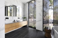 A modern bathroom features a sleek design with large floor-to-ceiling windows allowing plenty of natural light. The space includes two white vessel sinks with black fixtures on a wooden floating vanity. The walls and floors are designed with a blend of white marble patterns and dark tiles. A small, round wooden table is placed next to a bathtub adorned with potted plants and toiletries.