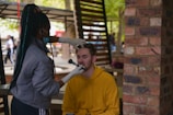 A person wearing gloves and a mask is applying makeup to another person sitting in an outdoor setting. The person receiving the makeup is wearing a yellow hoodie and has their eyes closed. There is a brick pillar and some blurred people in the background, indicating a casual environment.