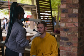 A person wearing gloves and a mask is applying makeup to another person sitting in an outdoor setting. The person receiving the makeup is wearing a yellow hoodie and has their eyes closed. There is a brick pillar and some blurred people in the background, indicating a casual environment.