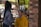 A person wearing gloves and a mask is applying makeup to another person sitting in an outdoor setting. The person receiving the makeup is wearing a yellow hoodie and has their eyes closed. There is a brick pillar and some blurred people in the background, indicating a casual environment.