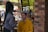 A person wearing gloves and a mask is applying makeup to another person sitting in an outdoor setting. The person receiving the makeup is wearing a yellow hoodie and has their eyes closed. There is a brick pillar and some blurred people in the background, indicating a casual environment.