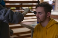A person with closed eyes is receiving makeup applied with a brush by another individual wearing gloves. The person receiving the makeup is wearing a yellow hoodie and has short brown hair. The background consists of blurred interior elements, suggesting an indoor setting.