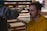 A person with closed eyes is receiving makeup applied with a brush by another individual wearing gloves. The person receiving the makeup is wearing a yellow hoodie and has short brown hair. The background consists of blurred interior elements, suggesting an indoor setting.