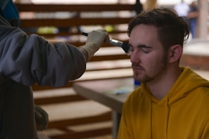 A person with closed eyes is receiving makeup applied with a brush by another individual wearing gloves. The person receiving the makeup is wearing a yellow hoodie and has short brown hair. The background consists of blurred interior elements, suggesting an indoor setting.