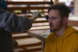 A person with closed eyes is receiving makeup applied with a brush by another individual wearing gloves. The person receiving the makeup is wearing a yellow hoodie and has short brown hair. The background consists of blurred interior elements, suggesting an indoor setting.