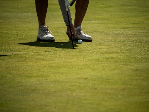 A person wearing white sneakers is placing a golf ball on a grassy field beside a golf club. The image focuses on the lower legs and feet of the individual, with a clear view of the grass and the shadow cast by the objects.