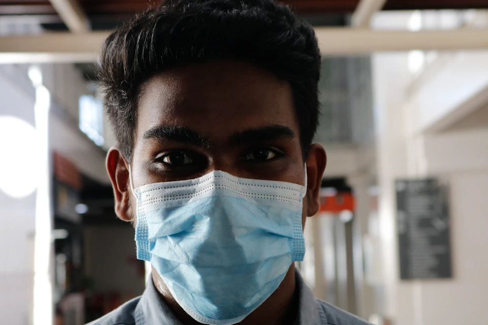 The image shows a young man wearing a blue surgical mask with white trim, standing against a plain white wall and a window. The man's face is the main focus of the image, which captures his expression and features prominently.