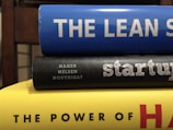 A stack of three books with colorful spines. The top book is blue with the title 'THE LEAN S'. The middle book has a black cover with the word 'startup' written on it and authors listed underneath. The bottom book is yellow, partially showing the title 'THE POWER OF H'. The books are stacked horizontally in a close-up view.