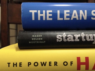 A stack of three books with colorful spines. The top book is blue with the title 'THE LEAN S'. The middle book has a black cover with the word 'startup' written on it and authors listed underneath. The bottom book is yellow, partially showing the title 'THE POWER OF H'. The books are stacked horizontally in a close-up view.