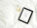 A white bed sheet serves as the backdrop for a bottle of Chanel No. 5 perfume and a framed picture. The frame contains an inspirational quote, 'Keep your heels, head & standards high,' in black text on a white background, surrounded by a black border.