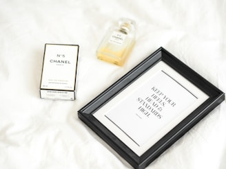 A bottle of Chanel No. 5 perfume is displayed alongside its box on a white surface. Next to it is a black framed quote that reads 'Keep your heels, head, and standards high.' The composition is minimalist and elegant.