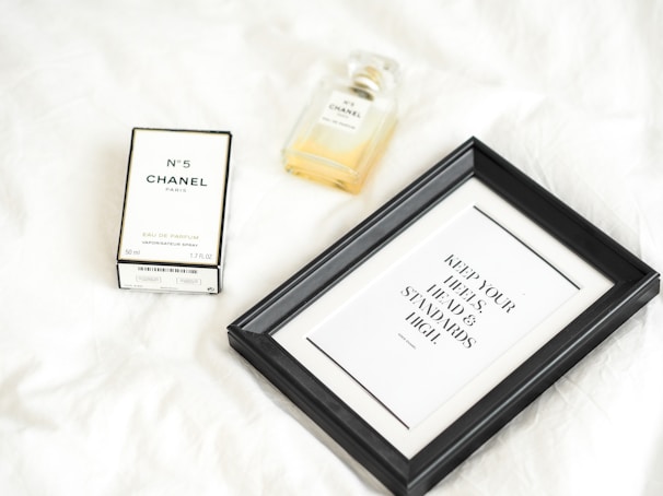 A bottle of Chanel No. 5 perfume is displayed alongside its box on a white surface. Next to it is a black framed quote that reads 'Keep your heels, head, and standards high.' The composition is minimalist and elegant.
