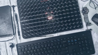 A workspace setup featuring a textured laptop placed on top of scattered papers. A spiral notebook, pen, wristwatch, and wireless earbud are arranged around it. A keyboard and wireless mouse are positioned below, all set against a backdrop of schematics and notes.
