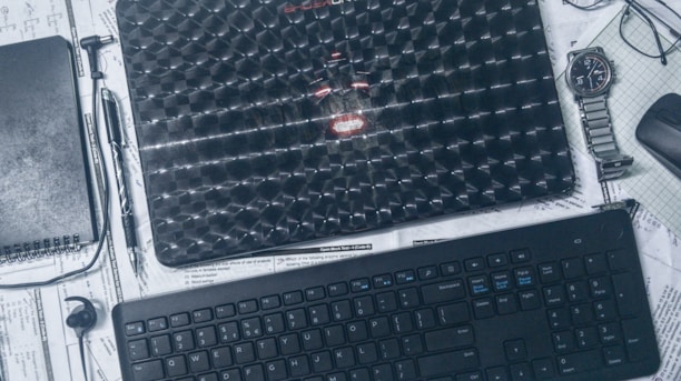 A workspace setup featuring a textured laptop placed on top of scattered papers. A spiral notebook, pen, wristwatch, and wireless earbud are arranged around it. A keyboard and wireless mouse are positioned below, all set against a backdrop of schematics and notes.