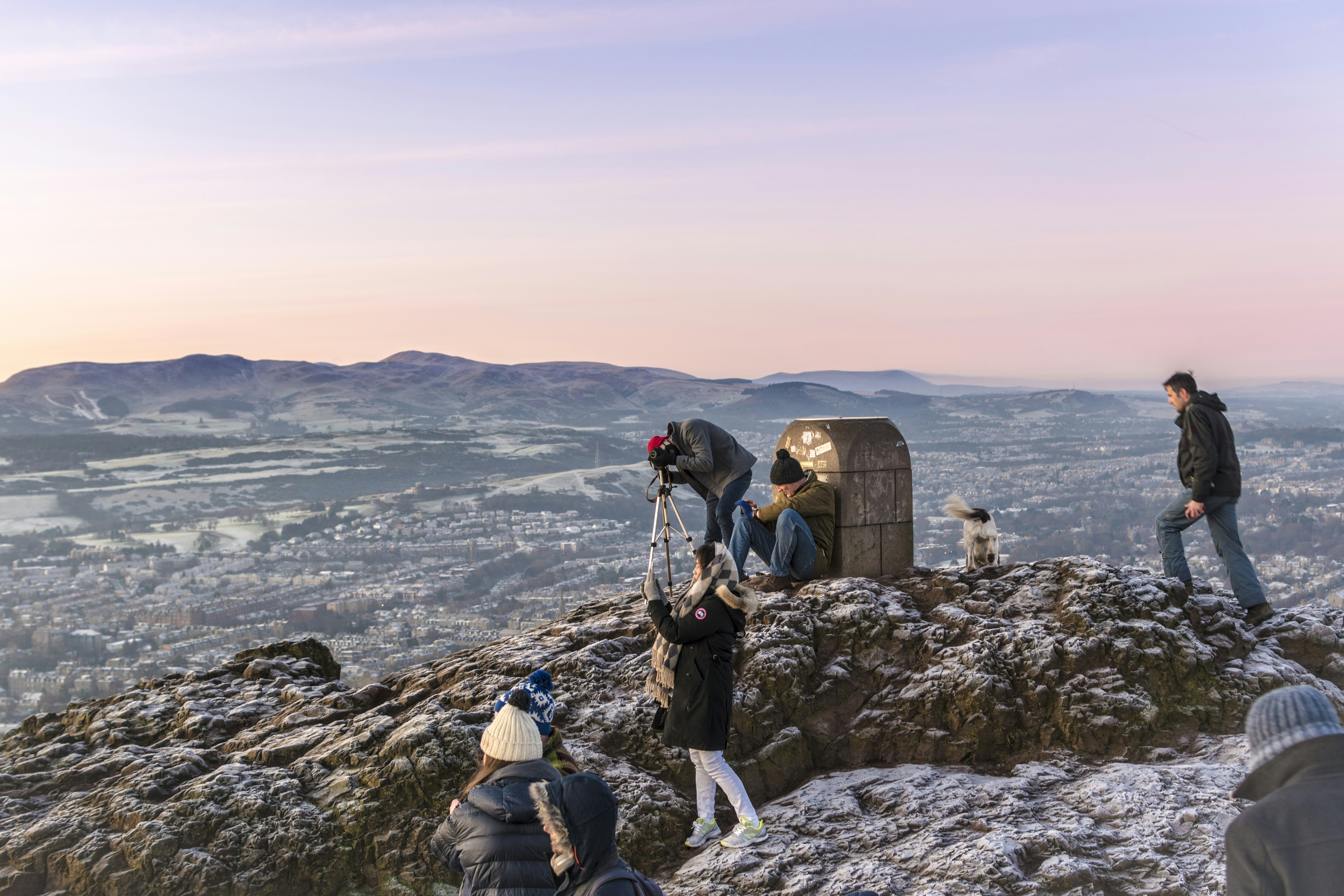 A dog and walkers on the frosty summit of Arthur's Seat at dawn, with the city and the Pentland Hills behind