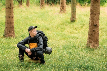 A person is crouching on a grassy forest floor surrounded by trees. They are wearing a black cap and a jacket with a yellow chest panel, and have a large backpack with a rolled-up sleeping mat attached.