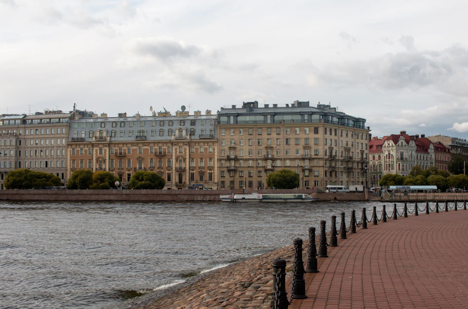 The image depicts a serene cityscape with a river running through it, flanked by buildings on both sides. The buildings vary in height and architectural style, creating a diverse skyline that reflects the city's history and character. The perspective is from across the river, providing a panoramic view of the urban landscape. The sky above is overcast, casting a soft light over the scene and enhancing the colors of the buildings.
