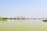 An industrial area with multiple smokestacks is reflected on the calm surface of a large body of water. The sky is clear and blue, and sparse green foliage is visible on the left side, adding a natural element to the otherwise industrial scene.