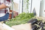A selection of fresh vegetables, including eggplants, herbs, and greens, are displayed in a wooden crate at an indoor market or store. The area appears organized with leafy greens arranged neatly. A person wearing a plaid shirt stands nearby, possibly tending to the display or shopping.