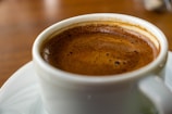 A close-up view of a steaming cup of coffee with a rich, dark crema on top, served in a white cup placed on a saucer. The background is softly blurred, emphasizing the coffee.