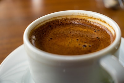 A close-up view of a steaming cup of coffee with a rich, dark crema on top, served in a white cup placed on a saucer. The background is softly blurred, emphasizing the coffee.