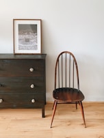 A minimalist interior featuring a dark wooden chair with an elegant, curved backrest to the right of a dark wooden dresser. The dresser has three drawers with ornate knobs, and a framed photo is placed on top, showcasing a large gathering of people. The setting is simple yet stylish, with light wooden flooring contrasting against the darker furniture.