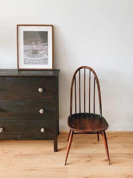 A minimalist interior featuring a dark wooden chair with an elegant, curved backrest to the right of a dark wooden dresser. The dresser has three drawers with ornate knobs, and a framed photo is placed on top, showcasing a large gathering of people. The setting is simple yet stylish, with light wooden flooring contrasting against the darker furniture.
