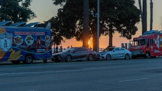 A vibrant street scene captures a pair of food trucks parked along a road near the ocean as the sun sets in the background. The trucks, painted in bright blue and red, are positioned in front of a couple of parked cars. Tall palm trees and a gathering of people silhouetted by the sunset complete the coastal atmosphere.