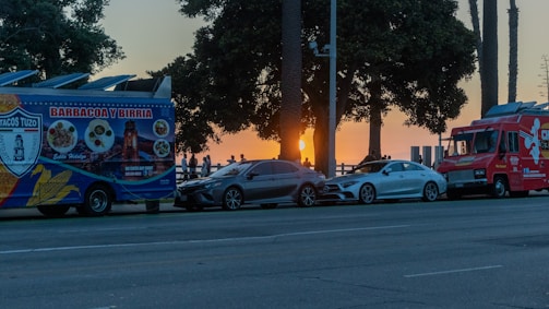 A vibrant street scene captures a pair of food trucks parked along a road near the ocean as the sun sets in the background. The trucks, painted in bright blue and red, are positioned in front of a couple of parked cars. Tall palm trees and a gathering of people silhouetted by the sunset complete the coastal atmosphere.