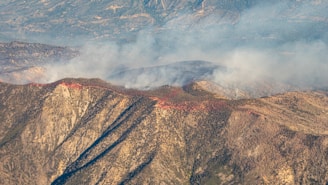 Aerial view of a mountainous landscape with a fire burning along the ridgeline. Smoke billows into the air, creating a hazy effect over the scene. The terrain is rugged and rocky, with variations in elevation and sparse vegetation.