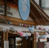 A quaint café shop with a wooden exterior is adorned with colorful string lights shaped like spheres, in pastel shades of pink, blue, and gray. A large circular sign with the words 'Café Shop' is prominently displayed above the entrance.