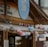 A quaint café shop with a wooden exterior is adorned with colorful string lights shaped like spheres, in pastel shades of pink, blue, and gray. A large circular sign with the words 'Café Shop' is prominently displayed above the entrance.