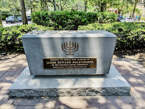 A memorial plaque set on a large stone block, surrounded by trees and greenery. The plaque contains a menorah symbol and text dedicating the site to James Edward Oglethorpe for the Savannah Jewish Community. Small stones are placed on top of the memorial.