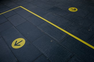 A dark stone pavement with two distinct yellow arrows and a yellow line, suggesting direction or guidance. The yellow elements are in sharp contrast with the dark color of the pavement.