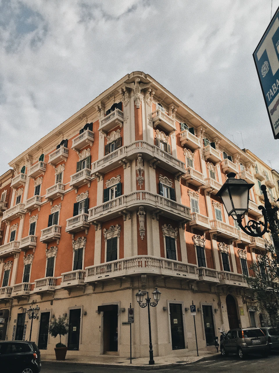 The image depicts a corner building with an orange facade, white trim, and balconies on each floor. The building is situated on a street corner, with cars parked along the sidewalk and trees visible in the background. A blue sign reading "TAB" can be seen on the right side of the frame.