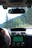 The interior of a car showing the driver's view with the steering wheel and dashboard visible. A person’s hand is on the steering wheel. The in-car navigation system is active and displaying a map on the screen. There are trees and greenery visible through the windshield, indicating an outdoor, possibly scenic, environment.