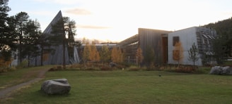 A modern building with angular, sloping roofs is surrounded by a green grassy area and trees with autumn foliage. The structure is made of wood and concrete, integrating with the natural landscape. A large rock is visible in the foreground, and the setting sun casts a warm glow over the scene.