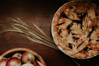 A freshly baked pie with a decorative, golden-brown crust sits on a dark wooden table. Next to the pie, there are some ears of wheat lying on the table. In the foreground, a bowl filled with red and green apples adds to the rustic setting.