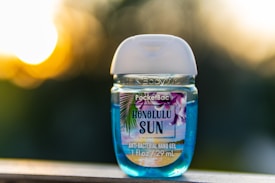 A small bottle of antibacterial hand gel labeled 'Honolulu Sun', featuring tropical imagery and a blue liquid inside. The background is an out-of-focus, warm-toned sunset.