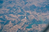 An aerial view of a landscape featuring a combination of agricultural fields, forested areas, winding roads, and a large body of water, possibly a lake or reservoir. The view captures a mosaic of different shades of green and brown, indicating varied land use and topography.