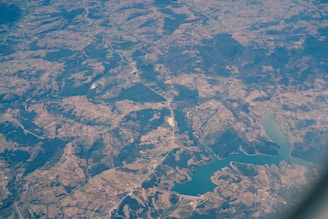 An aerial view of a landscape featuring a combination of agricultural fields, forested areas, winding roads, and a large body of water, possibly a lake or reservoir. The view captures a mosaic of different shades of green and brown, indicating varied land use and topography.