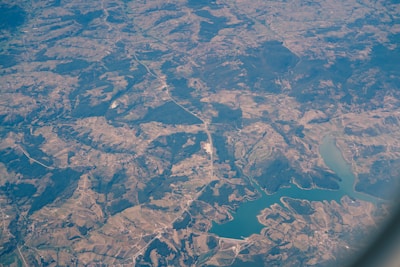An aerial view of a landscape featuring a combination of agricultural fields, forested areas, winding roads, and a large body of water, possibly a lake or reservoir. The view captures a mosaic of different shades of green and brown, indicating varied land use and topography.