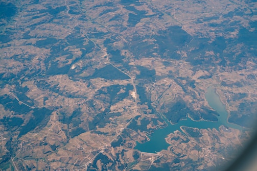 An aerial view of a landscape featuring a combination of agricultural fields, forested areas, winding roads, and a large body of water, possibly a lake or reservoir. The view captures a mosaic of different shades of green and brown, indicating varied land use and topography.