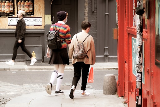 Two people are walking down an urban street. One of them is wearing a colorful striped sweater, black skirt, and knee-high socks, while the other wears a casual outfit with a patterned backpack. A pedestrian is seen walking in the opposite direction. The street is lined with shopfronts, including a red painted facade.
