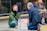 Two people engage in a social experiment involving eye contact, separated by a transparent acrylic sheet on a city sidewalk. One person is seated cross-legged, wearing a green jacket, while the other kneels across them wearing a suit. A sign between them reads 'Share One Minute Eye Contact'. A third person sits nearby, observing.