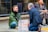 Two people engage in a social experiment involving eye contact, separated by a transparent acrylic sheet on a city sidewalk. One person is seated cross-legged, wearing a green jacket, while the other kneels across them wearing a suit. A sign between them reads 'Share One Minute Eye Contact'. A third person sits nearby, observing.