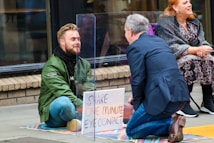 Two people engage in a social experiment involving eye contact, separated by a transparent acrylic sheet on a city sidewalk. One person is seated cross-legged, wearing a green jacket, while the other kneels across them wearing a suit. A sign between them reads 'Share One Minute Eye Contact'. A third person sits nearby, observing.
