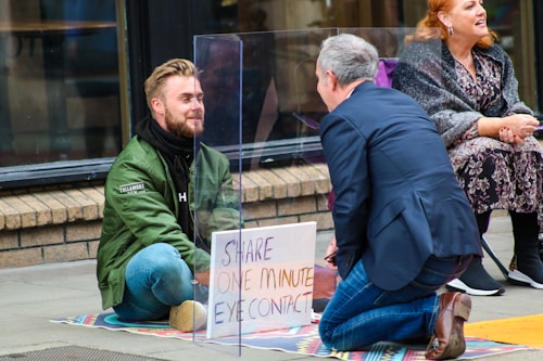 Two people engage in a social experiment involving eye contact, separated by a transparent acrylic sheet on a city sidewalk. One person is seated cross-legged, wearing a green jacket, while the other kneels across them wearing a suit. A sign between them reads 'Share One Minute Eye Contact'. A third person sits nearby, observing.
