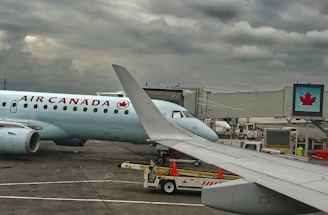 A plane from Air Canada is parked at an airport gate with a cloudy sky overhead. The airplane's wing is prominent in the foreground. Ground support vehicles and equipment are visible around the aircraft, along with a jet bridge leading to the plane. A digital sign with a maple leaf logo is displayed near the gate.