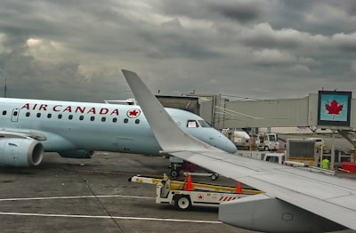 A plane from Air Canada is parked at an airport gate with a cloudy sky overhead. The airplane's wing is prominent in the foreground. Ground support vehicles and equipment are visible around the aircraft, along with a jet bridge leading to the plane. A digital sign with a maple leaf logo is displayed near the gate.